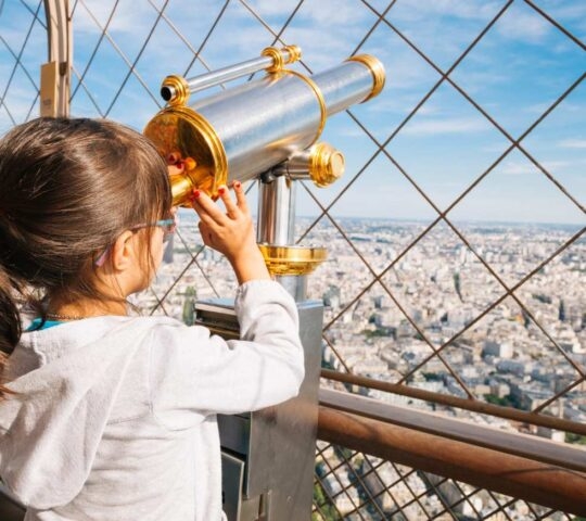 A young girl looks through a gold-colored telescope at the city of Paris from an observation deck.