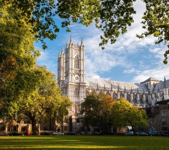 The exterior of Westminster Abbey is framed by lush green trees in a nearby park under a blue sky.