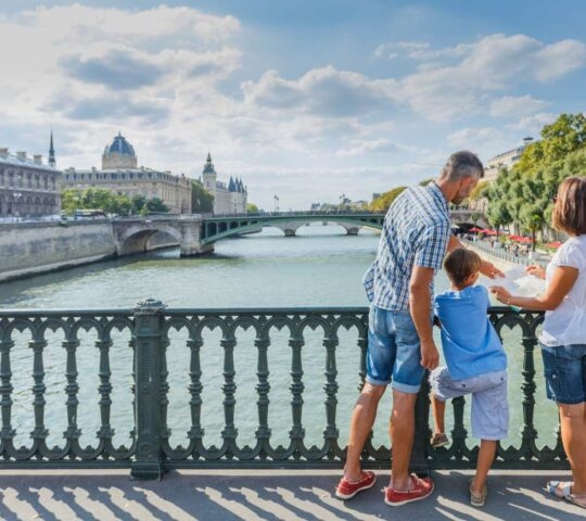 A family leans on a bridge railing while looking at a map and the river in a European city.