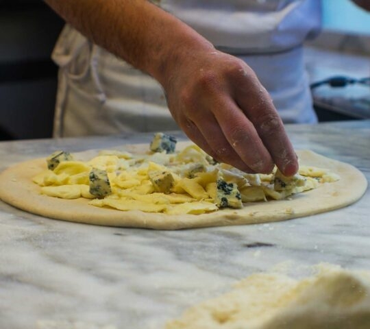Hands placing blue cheese onto raw pizza dough on a marble countertop.