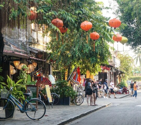 A street scene with many red lanterns hanging from trees above a parked bicycle and walking tourists.
