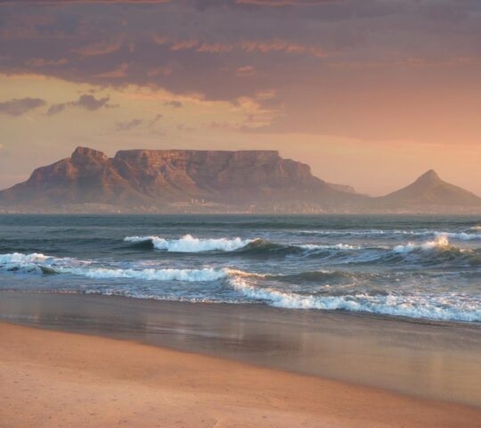 Ocean waves crashing on a beach with a large flat-topped mountain in the distance at sunset on a luxury Africa trip