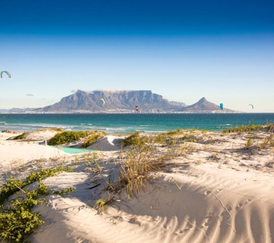 Sand dunes in the foreground overlook a blue bay with a large flat-topped mountain in the distant background.