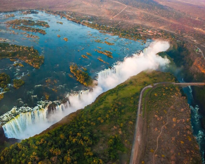 Aerial view of a wide waterfall with white mist rising into the air and lush green landscape along the riverbank.