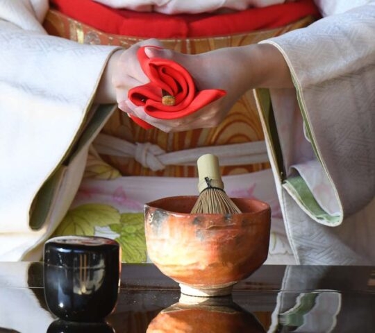 A close-up of a person in a kimono performing a Japanese tea ceremony, holding a folded red silk cloth above a matcha bowl and whisk.
