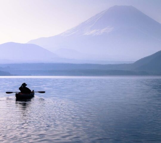 A person kayaks on a misty, calm lake in the foreground, with the snow-capped, blue-hued Mount Fuji visible in the distance.