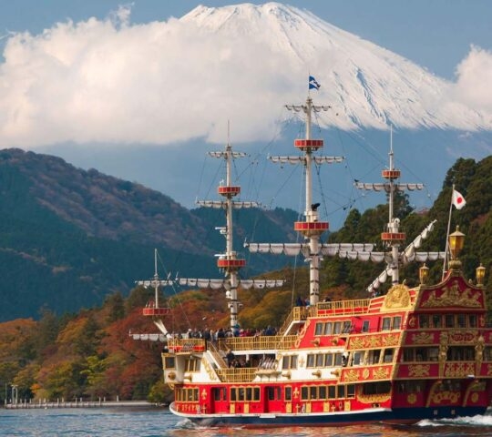 A large, ornate red and white tourist pirate ship sails on a lake, with the snow-capped Mount Fuji rising behind forested hills.
