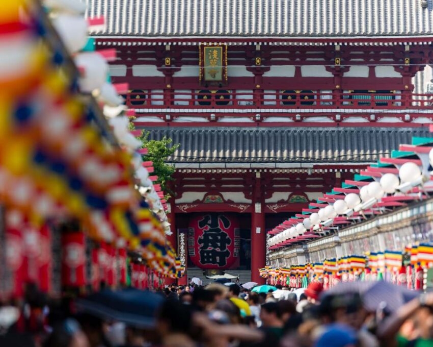 A crowded lane leading to a large red temple gate, lined with colorful banners, lanterns, and stalls, in Tokyo.