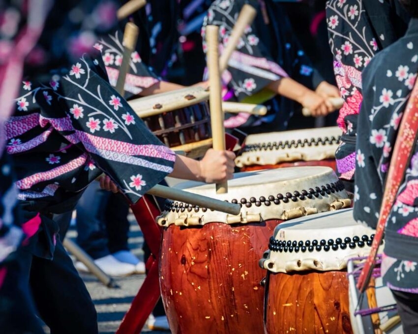 A close-up of people in traditional robes playing large Taiko drums with wooden sticks during a Japanese festival.