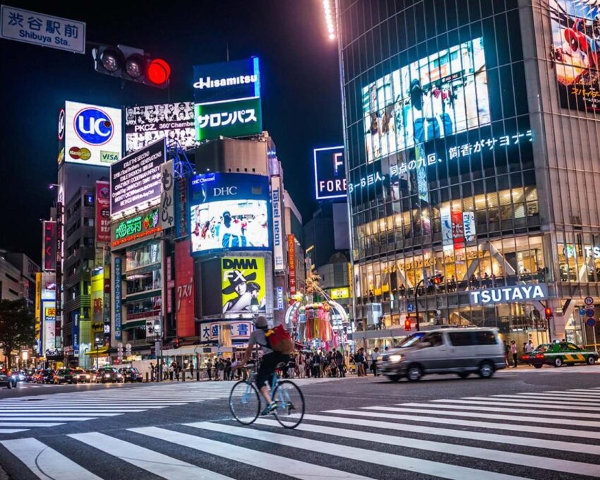 A nighttime view of Tokyo's Shibuya crossing with bright neon signs, skyscrapers, and a cyclist on the crosswalk.