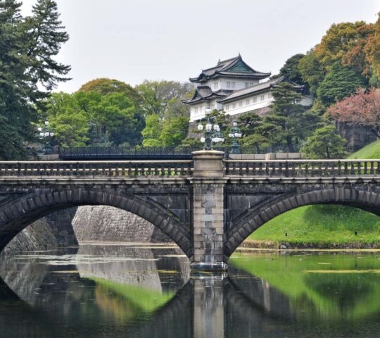 A stone arch bridge with reflections in a moat leading toward the traditional, white and green buildings of the Tokyo Imperial Palace.