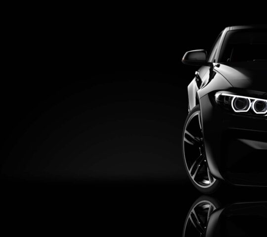 A dark, close-up, low-key view of a black sports car's front headlight and fender with a reflective surface.