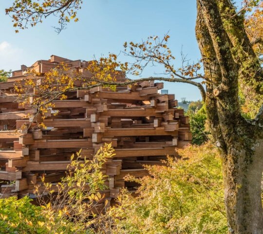 An abstract-looking, complex structure made of horizontally stacked brown wooden beams, surrounded by green and autumn trees.
