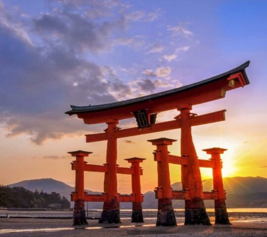 A large, red wooden torii gate standing in the water near a shore at sunset, with distant mountains.