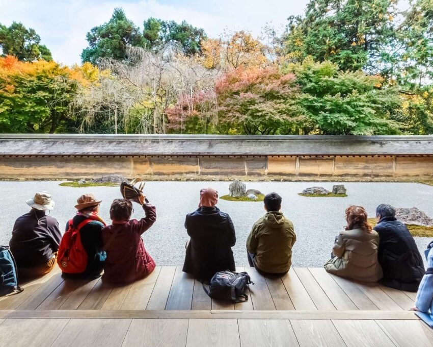 People sit on a wooden veranda, looking out over the stark white gravel and rock arrangement of the Ryoan-ji Temple rock garden.