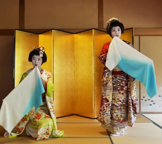 Two women dressed as maiko or geisha in colorful kimonos perform a dance holding silk cloths in front of a gold screen.