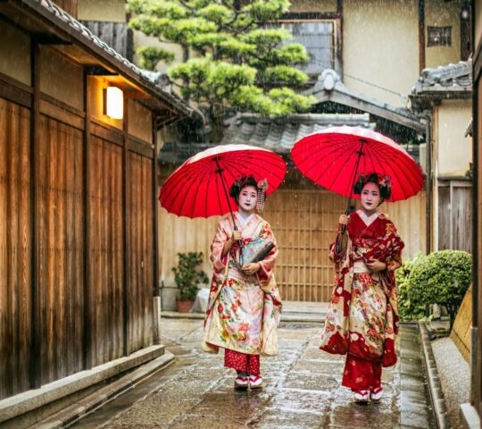 Two women dressed as maiko or geisha in kimonos walk down a historic Kyoto street, holding large red umbrellas in the rain.