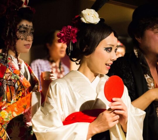 Close-up of three Japanese women, one in a white kimono holding a red mirror, one with black lace over her eyes, and one partially visible.
