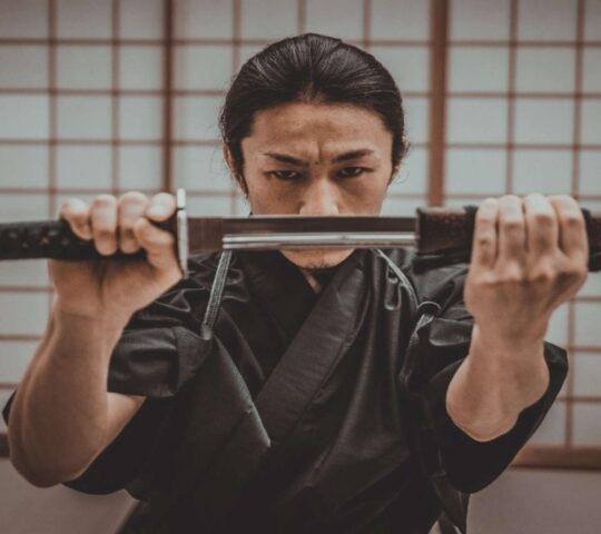 A man in a dark traditional Japanese outfit holding a sword horizontally, covering his lower face, with a traditional screen behind him.