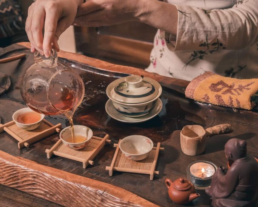 A close-up of hands pouring dark tea from a glass pitcher into small ceramic cups on a wooden tray, with a burning candle.