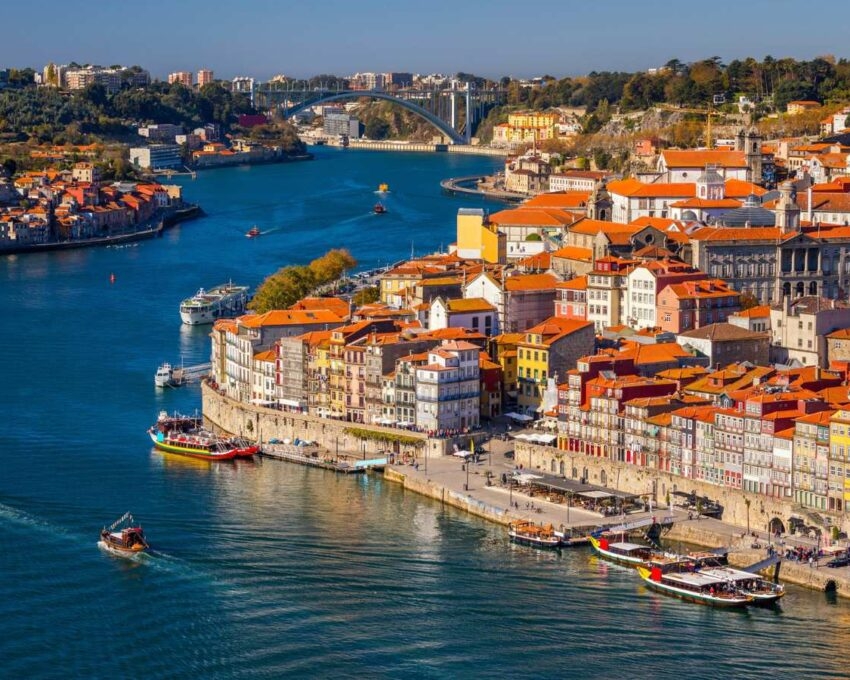 Aerial view of a city riverfront with densely packed red-roofed buildings and boats on the blue water.