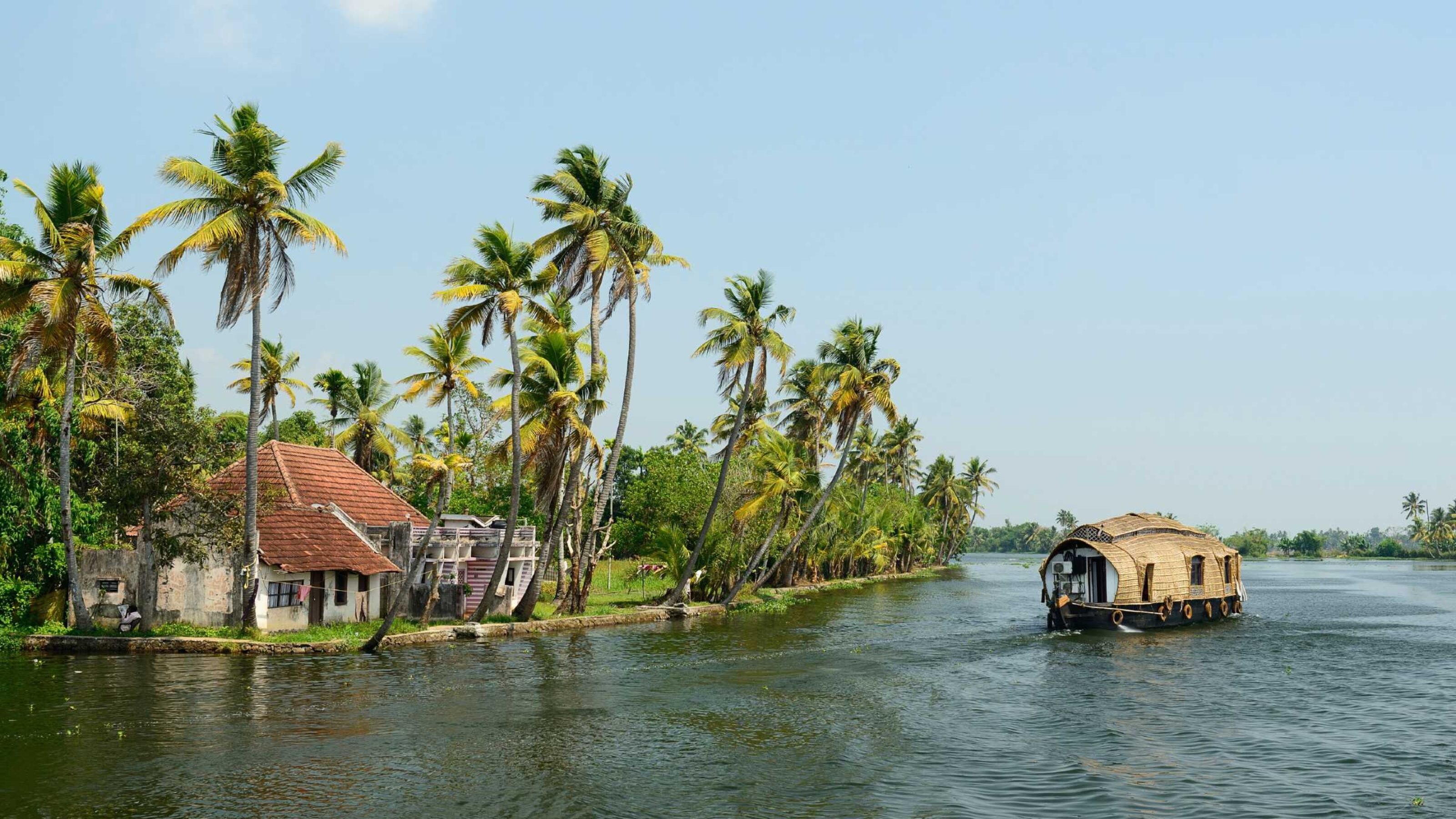 A wooden covered boat on a wide waterway in Kerala, with palm trees and a small red roofed building on the shore
