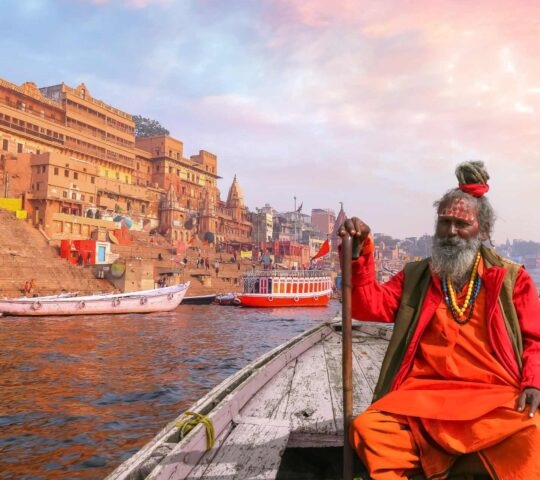 A man with grey hair and beard and wearing bright orange robes sitting in a boat on the River Ganges in the city of Varanasi