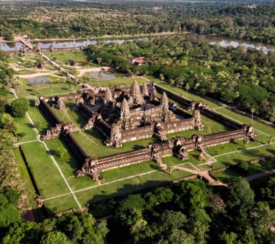 Aerial view of a large, ancient stone temple complex surrounded by lush green forests and clearings.