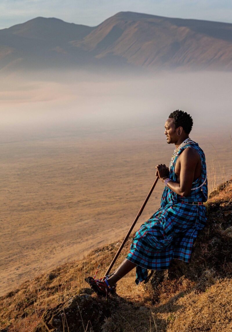A man in traditional blue clothing sits on a ridge overlooking a misty valley on luxury Ngorongoro Crater safari tours.