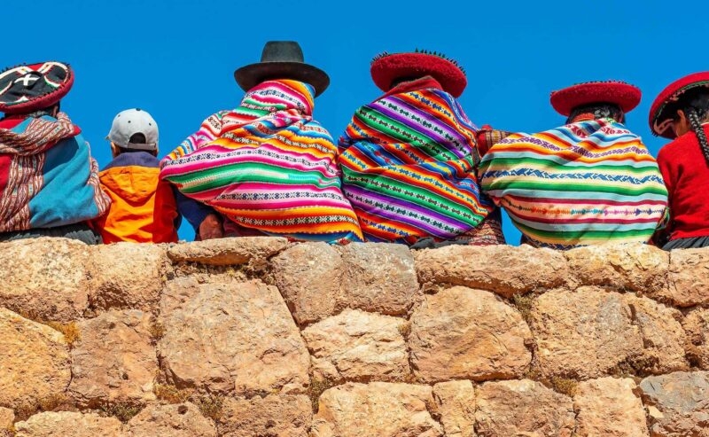 Panoramic photograph of Quechua indigenous women in traditional clothing with a boy sitting on an ancient Inca wall in Chinchero, Cusco Province, Peru.