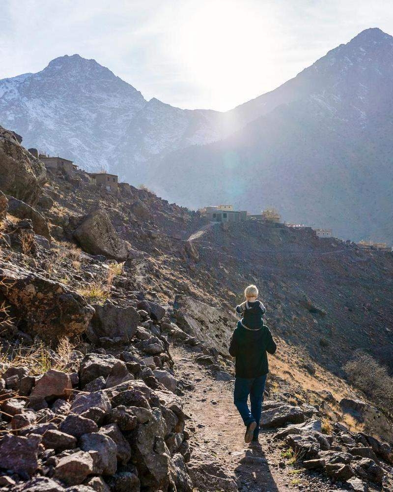 A hiker with a child on their shoulders walks along a steep, rocky path with snow-capped mountains in the background.