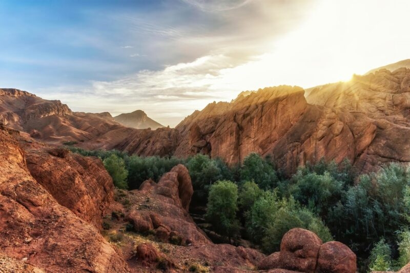A wide shot of a rocky canyon with lush green vegetation in the valley during a bright sunset.