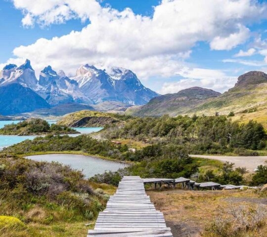Wooden plank path leading through a valley toward bright blue lakes and steep, snow-dusted granite mountains.