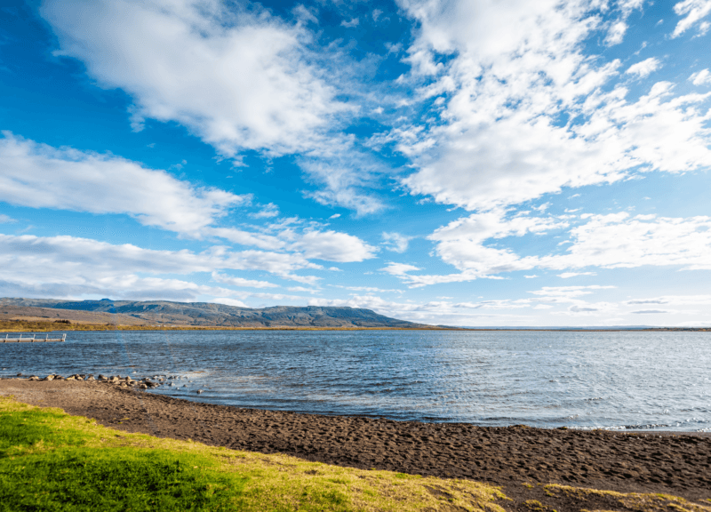 A scenic lake view from a luxury golden circle tours stop, featuring a dark sand shore and blue sky with white clouds.