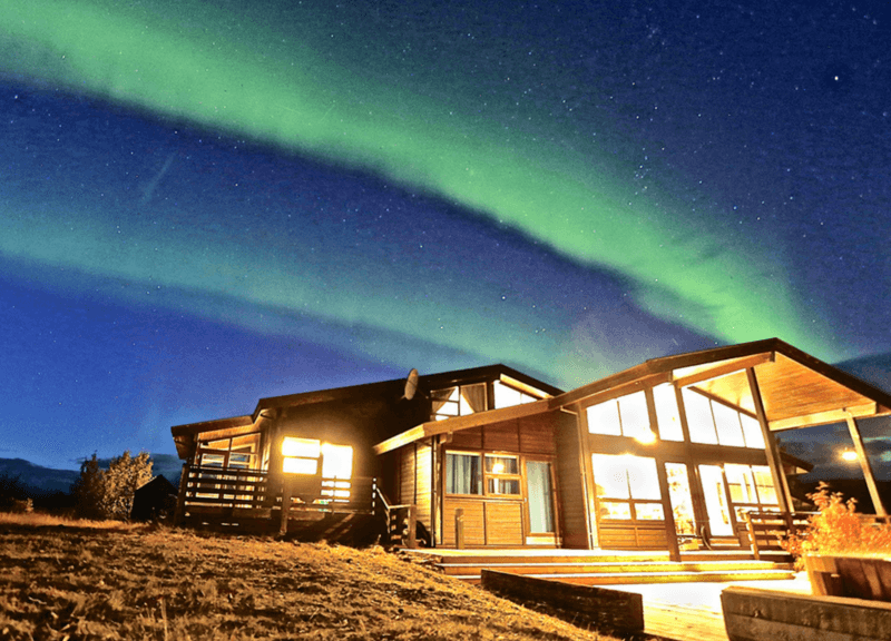 A luxury cabin with illuminated windows at night under a starry sky and green aurora borealis during luxury Golden Circle tours.