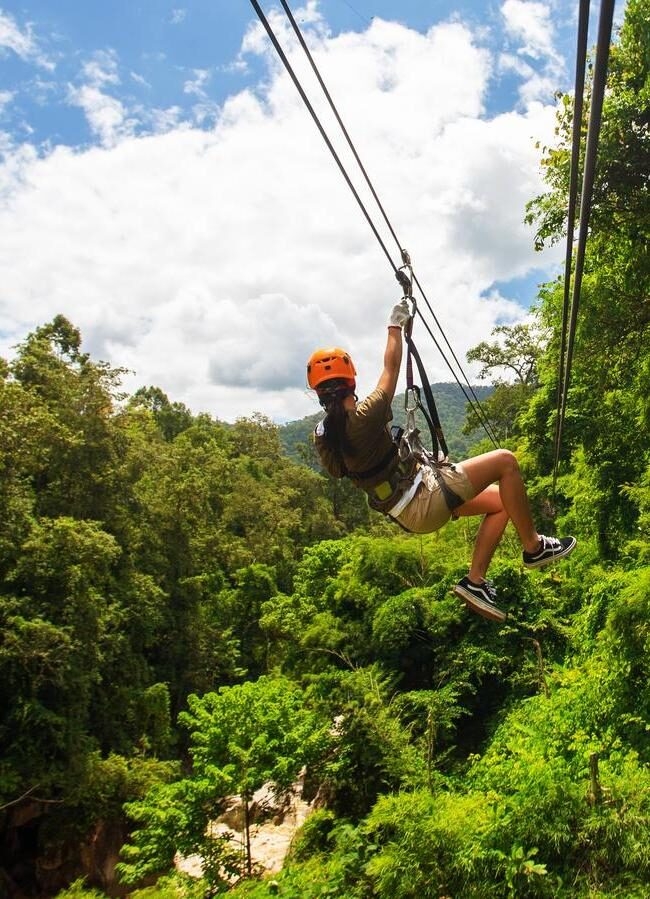 A person ziplining through a dense green tropical forest under a blue sky with white clouds.