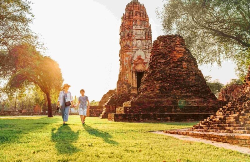 A woman and boy walking past ancient stone ruins and a tall weathered prang tower during a sunny afternoon.