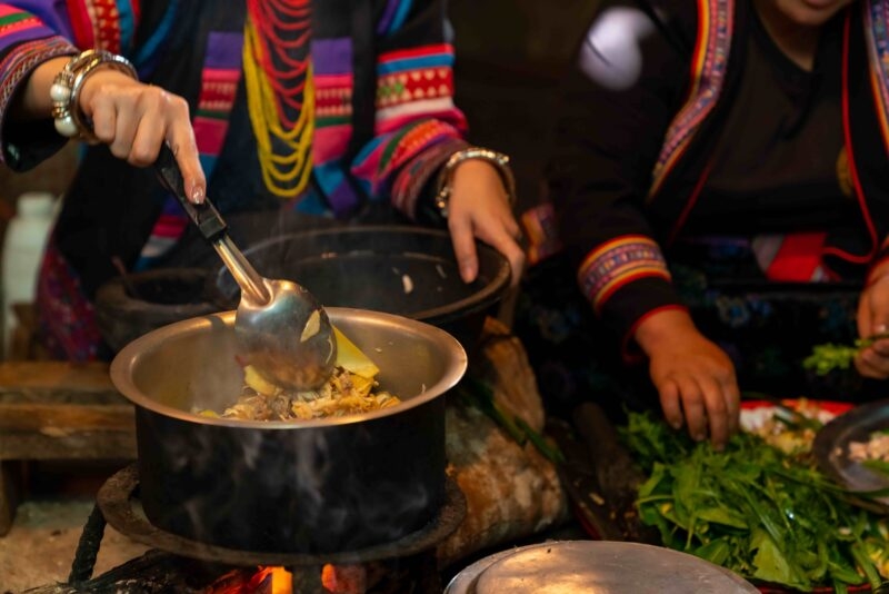 Close-up of hands stirring food in a metal pot on a stove, with people in colorful traditional ethnic dress.