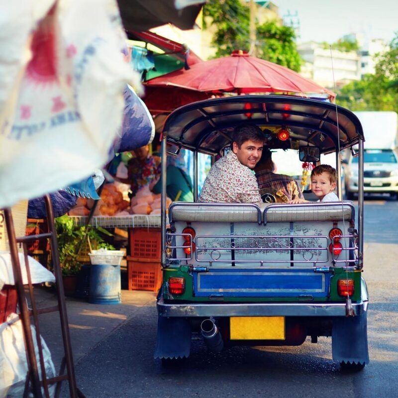 A man and a young boy looking back from the rear seat of a blue and green tuk-tuk on a city street.