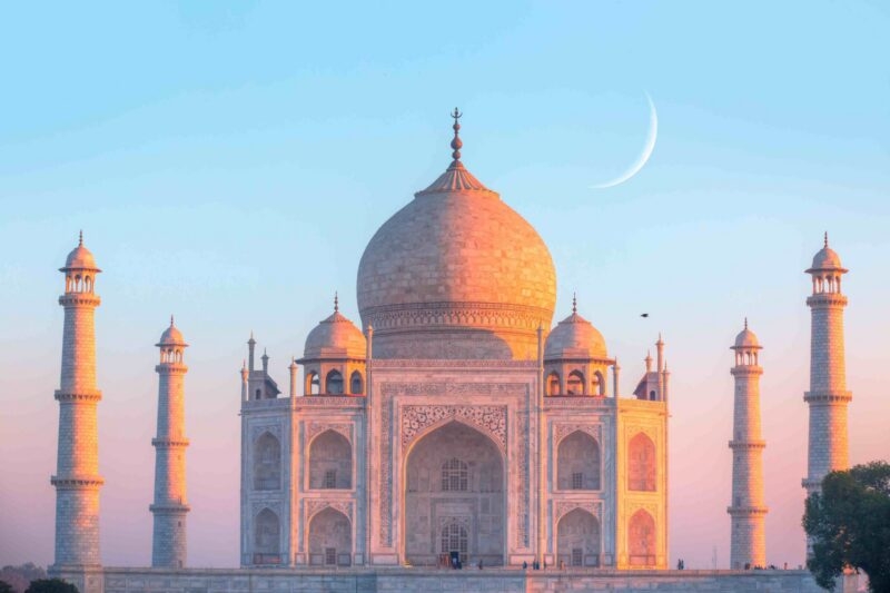 The Taj Mahal building and its minarets at sunset under a pale blue sky with a visible crescent moon.