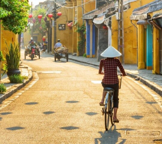 Vietnamese woman in traditional hat bicycling along in Hoi An