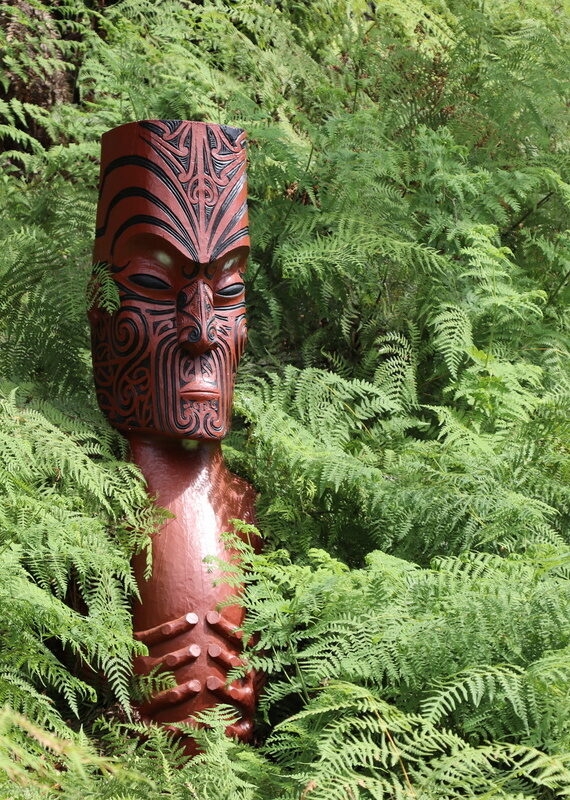 Sculpture of a Māori Totem, a carved wooden figure amongst thick fern foliage