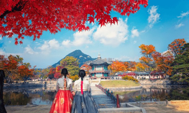 Two women in traditional Hanbok clothing stand with their backs to the camera, looking at a classic Korean pavilion across a pond in autumn.
