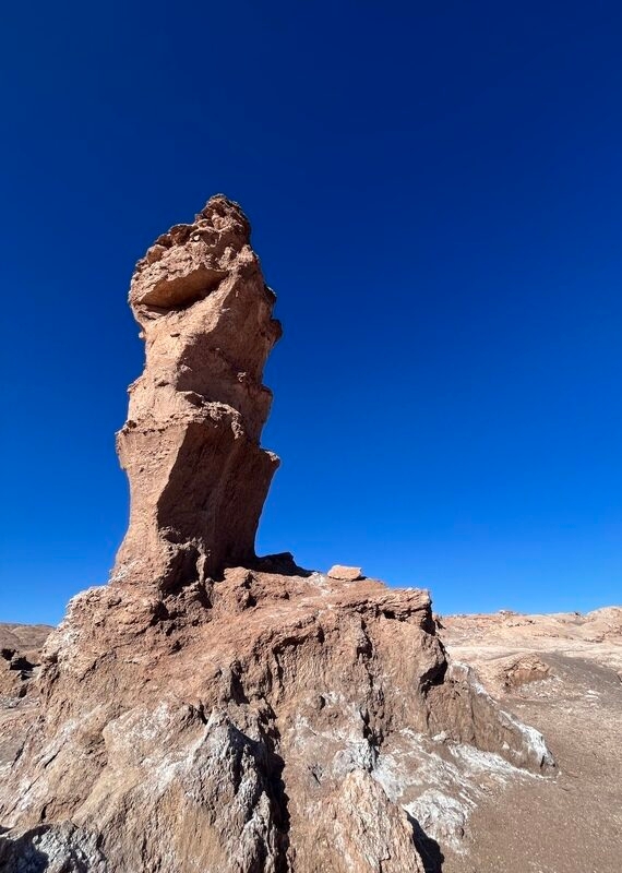 A tall, rugged rock formation stands against a clear blue sky, showcasing the arid landscape of a desert region.