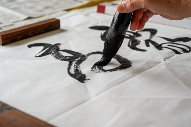 A hand holding a calligraphy brush painting black ink characters on white paper.