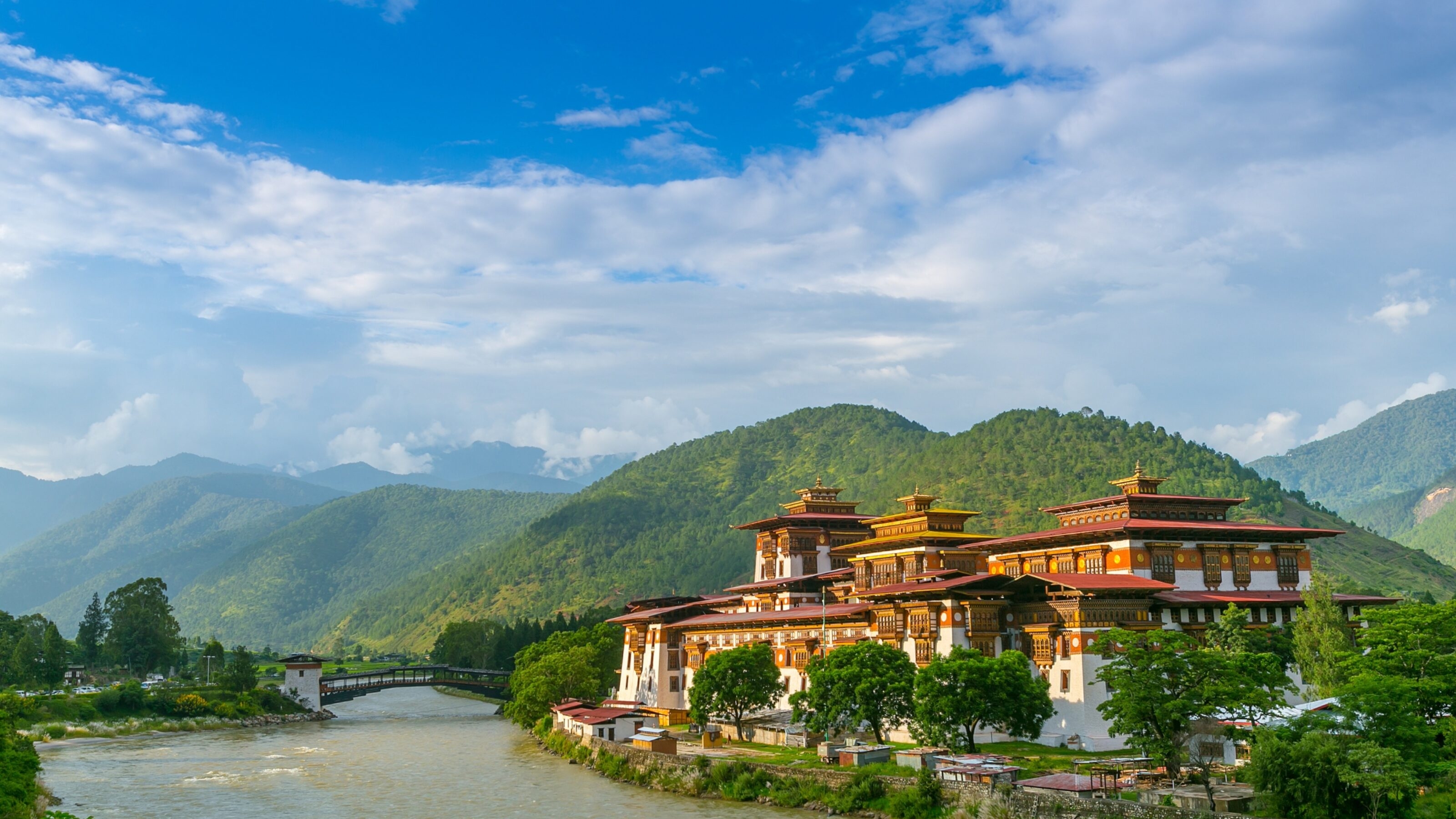 Traditional Bhutanese architecture style building with a river and bridge against a backdrop of green mountains and blue sky.