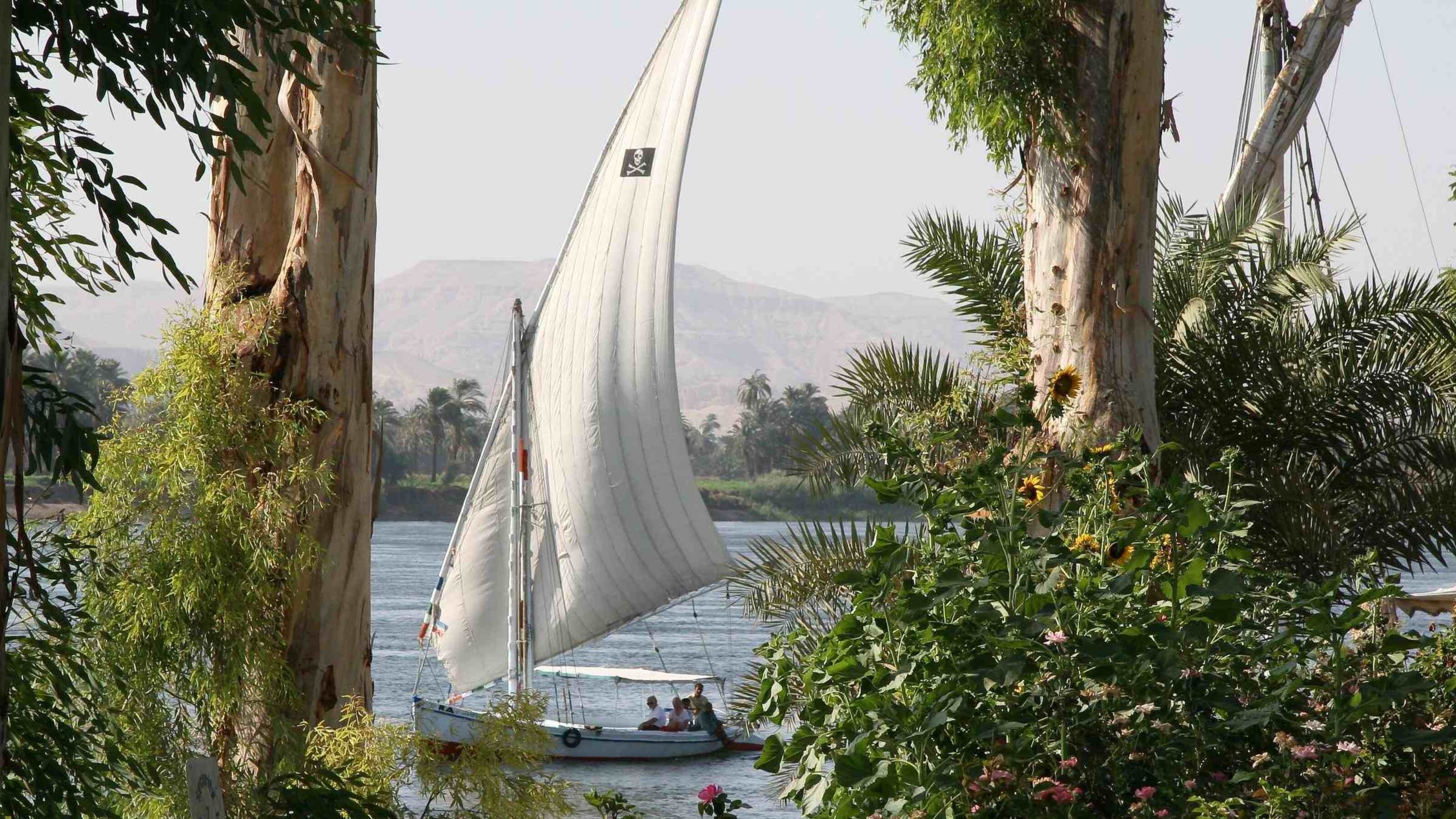 Sailboat on river with lush greenery and mountains in the background.