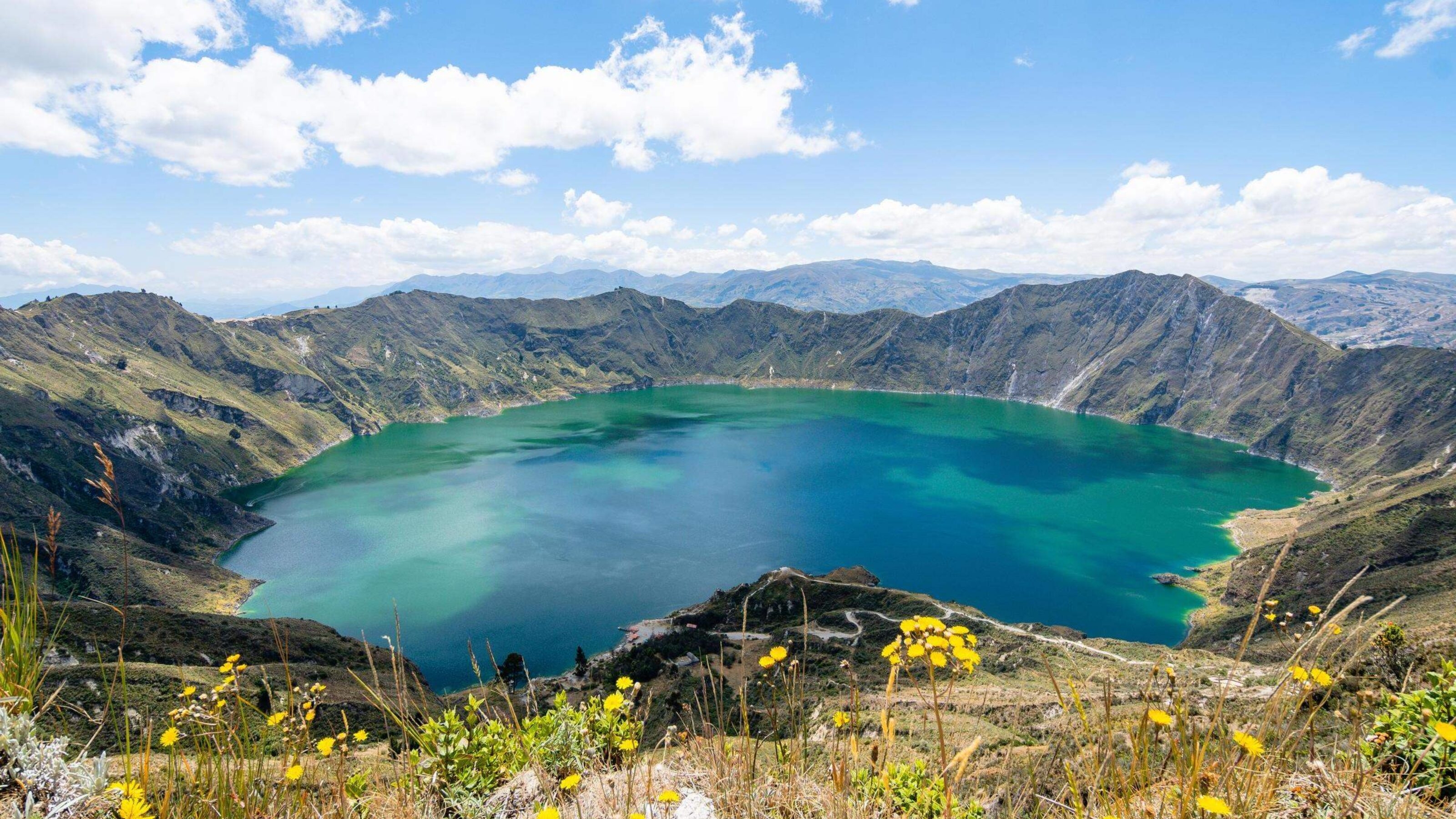 A panoramic view of a mountainous landscape with a vivid blue crater lake under a cloudy sky.