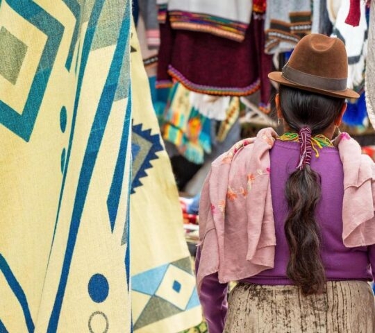 Indigenous Ecuadorian Otavalo woman in traditional clothing, hat and hairstyle on Otavalo local market with textile and fabric stalls, Ecuador.