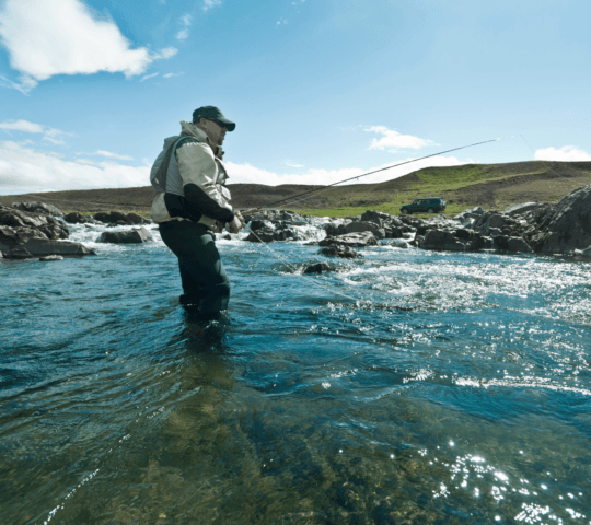 Person in waders fly-fishing in a sparkling river with rocky banks under a blue sky.